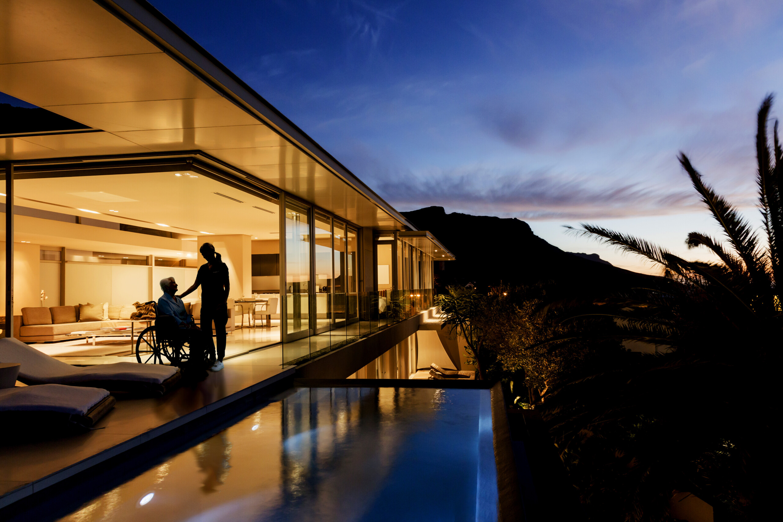 Modern glass-walled house at dusk; a man in a wheelchair is assisted by another person near a couch by the pool.