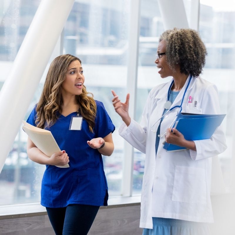Two healthcare professionals converse in a bright hospital hallway: a nurse in blue scrubs holding folders speaks with a doctor in a white coat and stethoscope.
