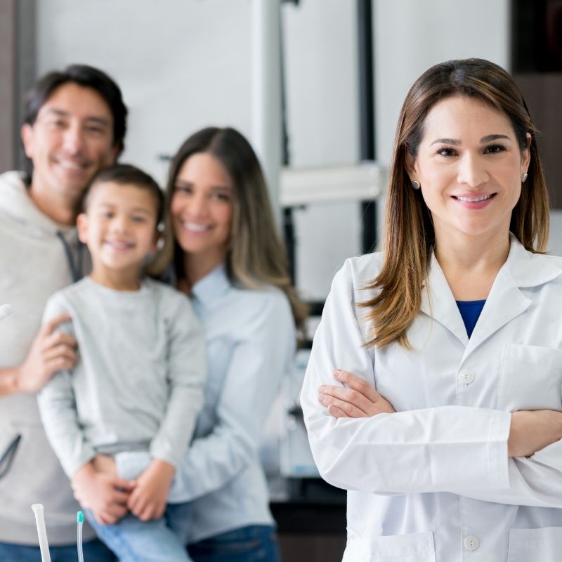 Smiling female doctor in a white coat with arms crossed, a happy family blurred in the background at a clinic.