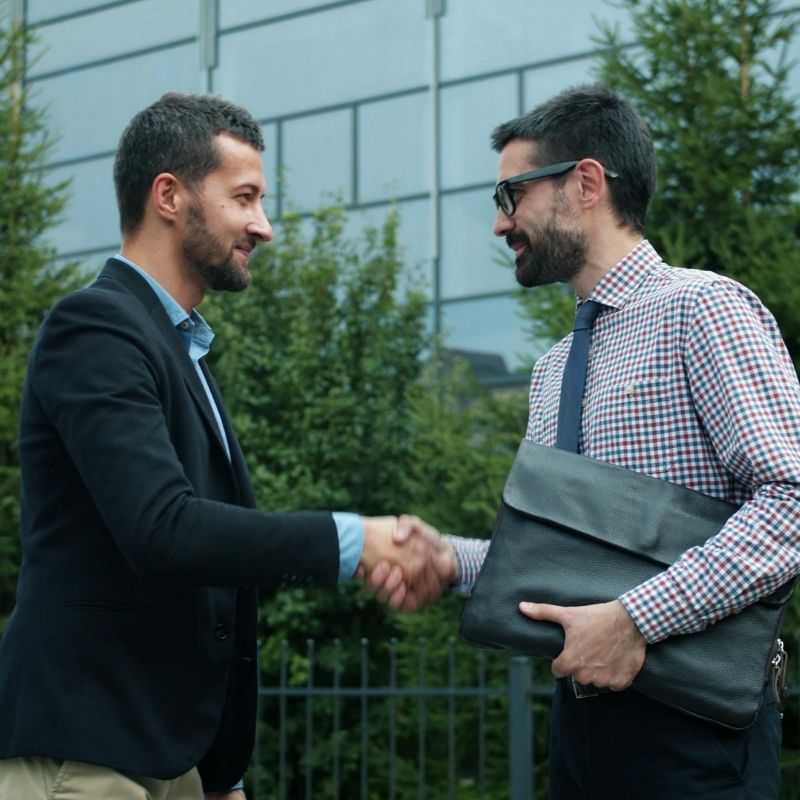 Two men in business attire shake hands outdoors, one holding a black leather portfolio and a gray laptop bag, in front of a glass building and greenery.
