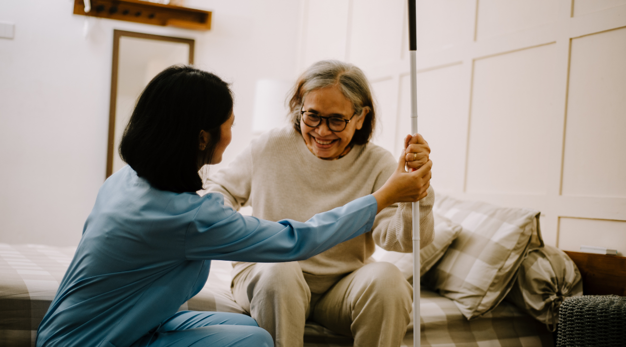 Caregiver in blue scrubs helps an elderly woman stand with a walking cane, both smiling at a bedside in a cozy room.